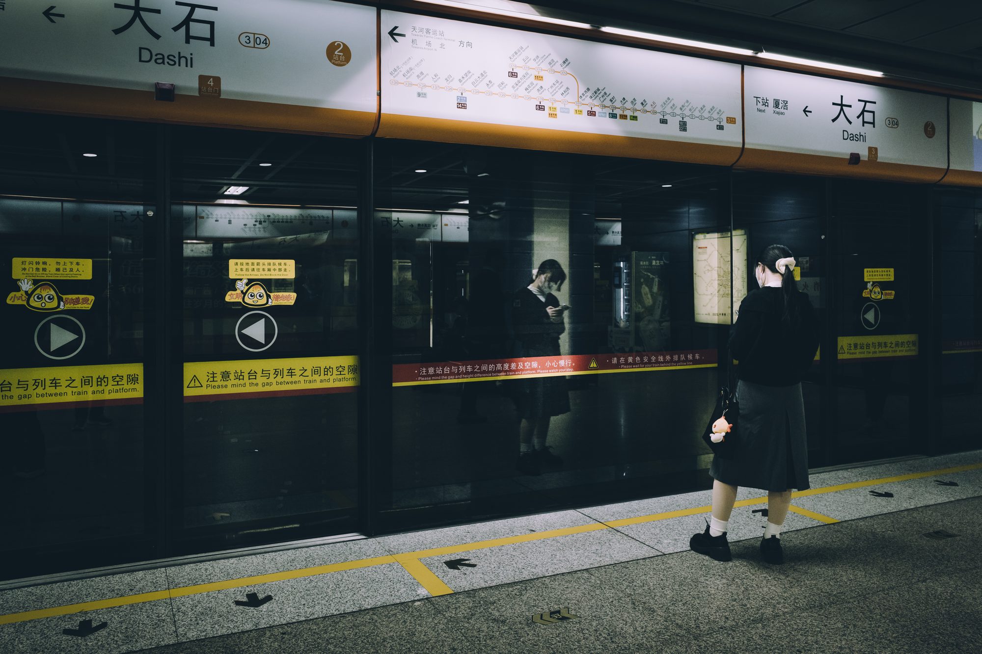 广州地铁大石站站台，一位穿长裙女子背对镜头独自等车，紧闭的玻璃屏蔽门后另一位乘客正在看手机
