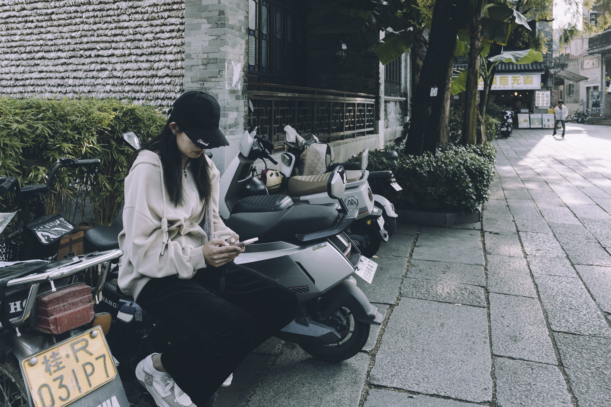 老城街边停了一排电瓶车，戴黑色棒球帽的女子斜坐在其中一辆车上低头看手机，背景是青砖巷子和挂着灯笼的商铺