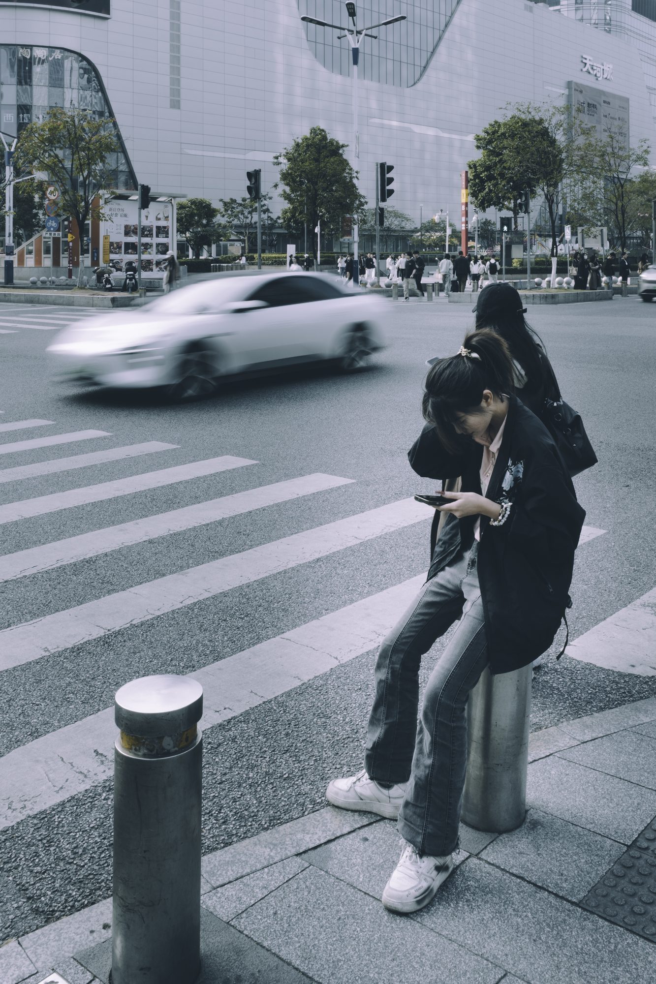 街头斑马线旁的金属路桩上，穿黑色外套的年轻女子坐着低头看手机，背景是模糊飞驰的白色汽车与车流