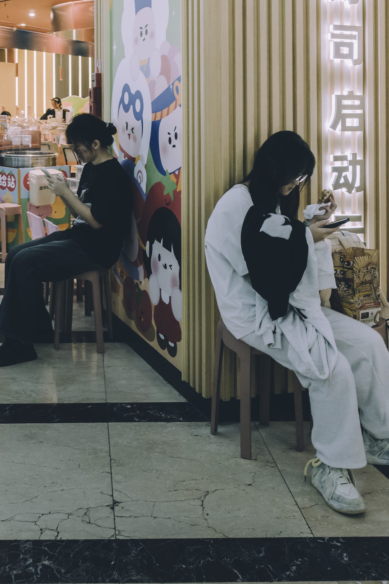 商场餐饮区里一面卡通角色墙画前，两位年轻女子分坐两个小凳上各自低头看手机，彼此并无交流