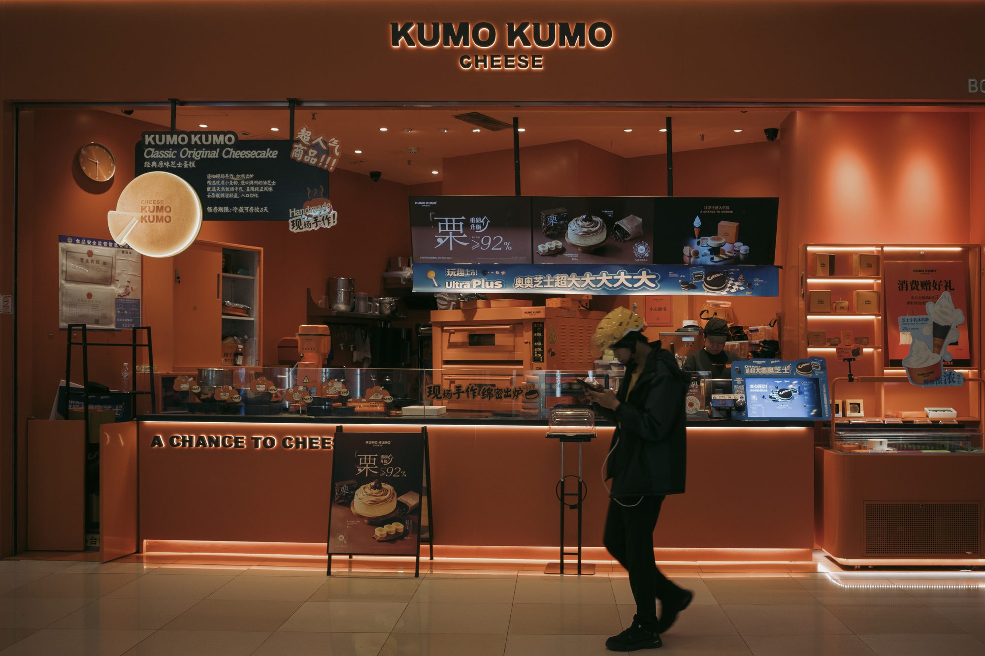 KUMO KUMO 芝士蛋糕店明亮的橙色橱窗前，戴黄色帽子穿黑色外套的人从店面走过，仍在低头看手机