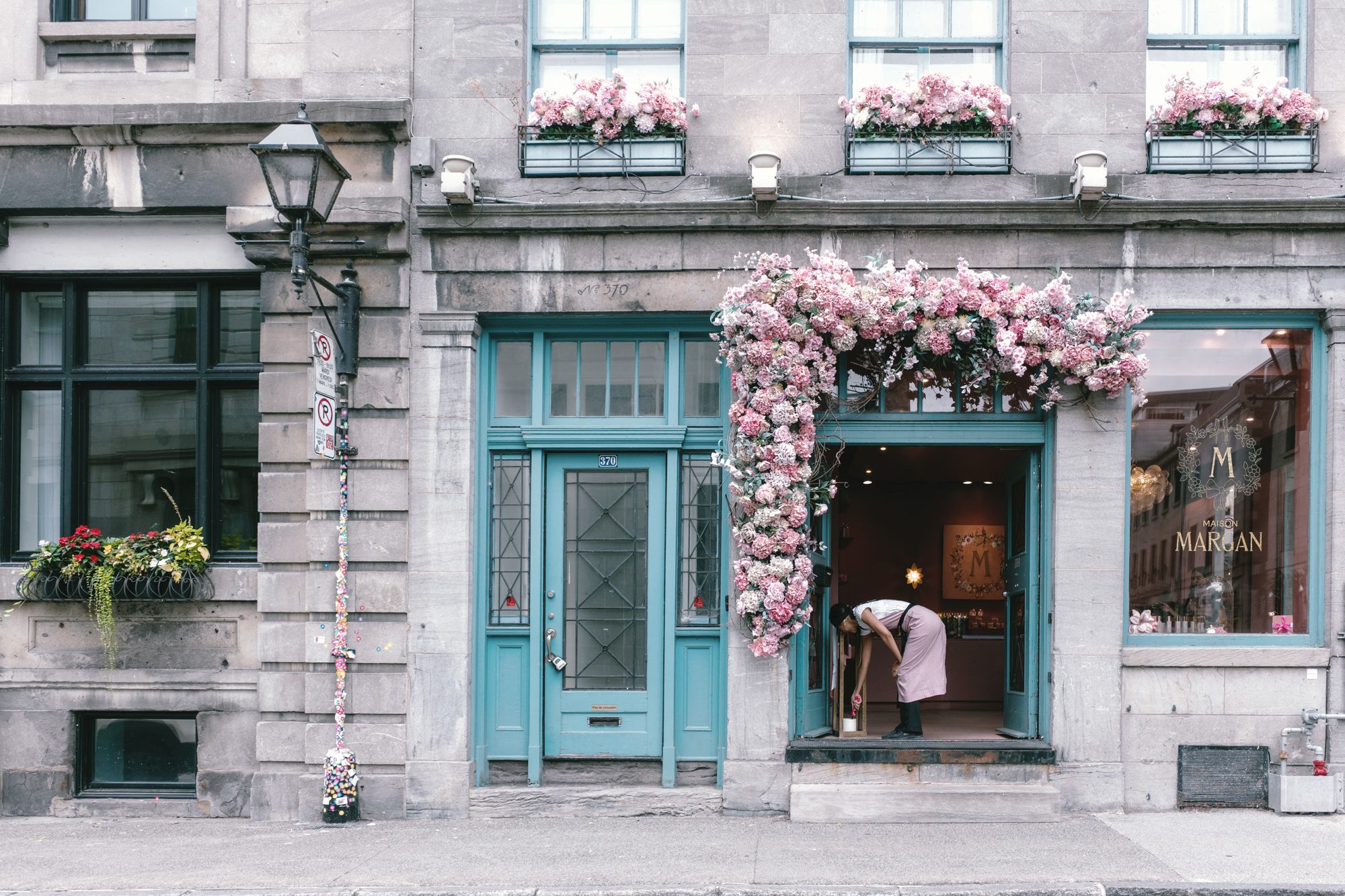 灰石外墙的「Maison Margan」店面，门框和窗框漆成青绿色，门口垂着大片粉色花饰，一名穿粉色围裙的店员正弯腰整理门口的东西