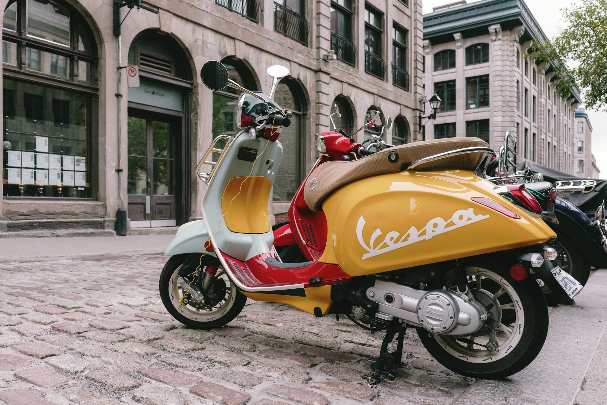 老城鹅卵石街边停着一辆红黄双色的 Vespa 踏板摩托车，背景是灰色石砌的商铺与空荡的街道