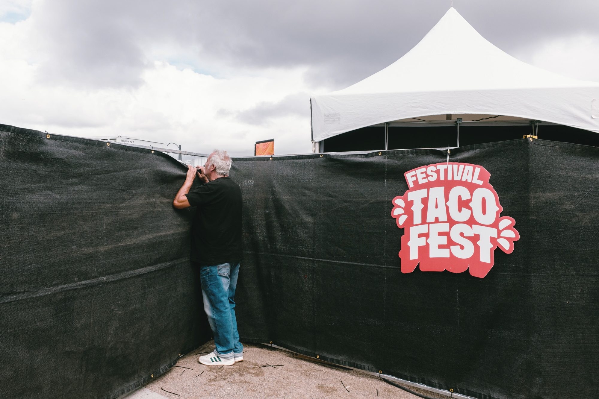 一名灰发男子踮脚扒着黑色围挡向内张望，围挡上挂着红色的「Festival Taco Fest」标志，后方是白色帐篷和厚重的云
